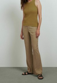 Haut sans manches tricoté vert avec ourlet côtelé, porté avec un pantalon beige évasé. Le modèle porte des sandales noires. Texture simple et lignes épurées.