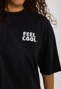 Schwarzes Baumwoll-T-Shirt mit kurzen Ärmeln, das auf der linken Brustseite eine weiße Grafik mit der Aufschrift "FEEL COOL" zeigt.