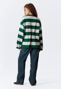 Zeleno-sivý pruhovaný dlhý rukáv s voľným strihom, skombinovaný s tmavomodrými širokými nohavicami, na jasnom bielom pozadí.