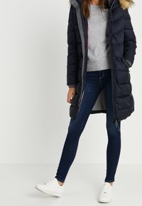 Marinblå puffjacka med fake päls på huvan, fickor med dragkedja och grå foder. Bärs över en grå tröja och mörkblå jeans med vita sneakers.