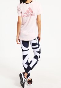 Žena oblečená v světle růžovém tričku Adidas, černých a bílých legínách s vzorem a černých a růžových teniskách Adidas, stojící zády.
