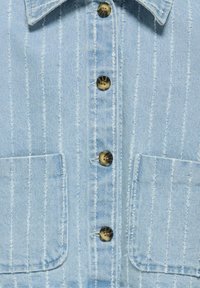 Svetlo modra denim srajca z navpičnimi belimi črtami, petimi mesingastimi gumbi spredaj in dvema zamaščenima žepoma. Prikazani so obrabljeni detajli.