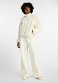 Fleece trainingspak in crème met lichtbeige accenten. De jas met rits heeft een hoge kraag; de broek is wijd en heeft een ontspannen pasvorm.