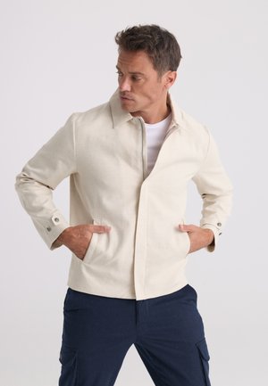 Homme portant une veste zippée beige clair sur une chemise blanche, les mains dans les poches de la veste, regardant vers sa gauche.