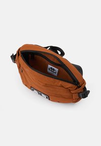 Oranje stoffen schoudertas met een ritsluiting, zwarte accenten en een label aan de binnenkant. Heeft een compacte, rechthoekige vorm.