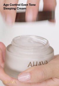 Pot en verre de crème avec une surface texturée et un couvercle en métal argenté, contenant une crème de nuit lisse et blanche. Logo "AHAVA" en doré.