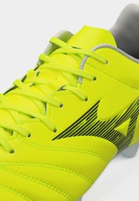 Φωτεινό κίτρινο παπούτσι ποδοσφαίρου με κορδόνια νέον κίτρινου χρώματος, γκρι εσωτερική επένδυση και μαύρο λογότυπο με ρίγες στη πλευρά.