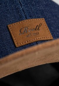 Gorra de mezclilla con un parche de cuero marrón con texto en relieve. La gorra tiene un color azul oscuro con una tela texturizada y detalles de costura.