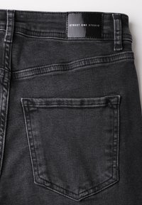 Vaqueros de mezclilla negra con un bolsillo trasero visible y una etiqueta brillante en la cinturilla marcada como "STREET ONE STUDIO". Detalles de costura distintivos.