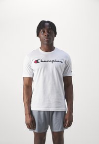 Camiseta blanca de manga corta con un logo "Champion" en negro y rojo. Modelo llevando pantalones cortos de malla grises. Diseño sencillo con cuello redondo.