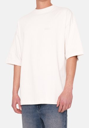 Personne portant un t-shirt blanc uni à manches courtes surdimensionné avec un col rond et un jean en denim bleu clair, debout devant un fond uni.