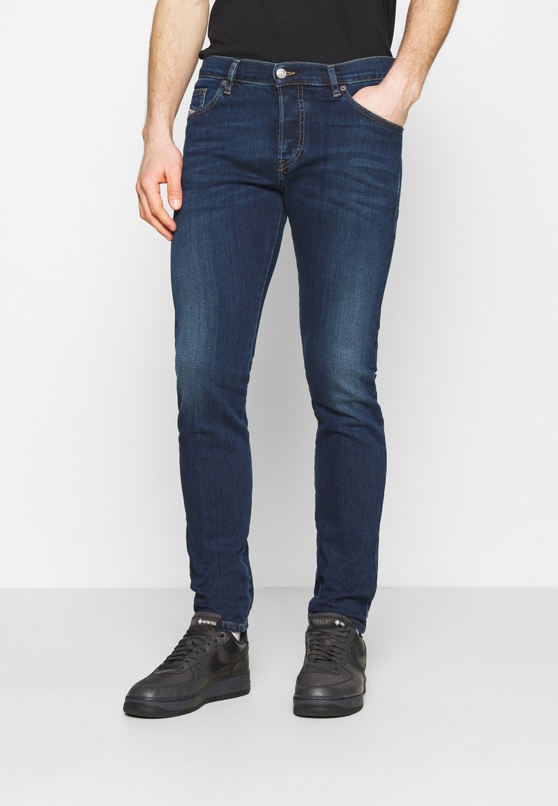 Dunkelblaue Slim-Fit-Denim-Jeans mit glatter Textur, fünf Taschen und kontrastierenden Nähten, kombiniert mit schwarzen Sneakern.