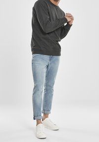 Uomo che indossa un felpa grigio scuro, jeans azzurri arrotolati e sneakers bianche, in piedi con le mani unite davanti.