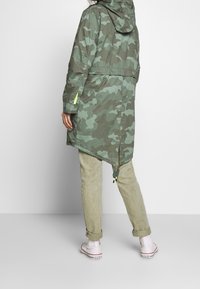 Green camo raincoat with a hood, featuring a longer back, adjustable drawstring hem, and contrasting yellow accents. Worn over light pants and sneakers.