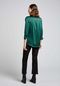 Blouse verte, légère, à manches longues avec un motif texturé, poignets roulés et ourlet arrondi, associée à un pantalon noir évasé et des bottes à talons larges.
