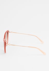 Occhiali da sole tonalità rossa con montatura quadrata, bracci in metallo dorato e dettagli a forma di cuore sulle cerniere. Finitura in plastica testurizzata.