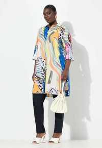 Blusa colorata e decorata con design di farfalle in blu, arancione e rosa, abbinata a pantaloni scuri e sandali con tacco bianchi. Sorreggendo una borsa color crema.