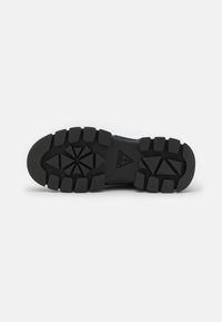 Semelle de botte en caoutchouc noir avec des rainures profondes et le logo triangulaire Guess au centre, présentée sur un fond blanc.