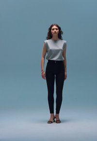 Γκρι αμάνικη μπλούζα, μαύρο ψηλόμεσο παντελόνι και μαύρα πέδιλα με τακούνι. Το μοντέλο στέκεται μπροστά σε ανοιχτό μπλε φόντο. Απλό, εφαρμοστό σχέδιο.