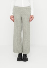 Personne portant un pantalon large en maille torsadée gris clair, un haut à manches longues blanc et des bottes noires à lacets épaisses, debout contre un fond blanc.
