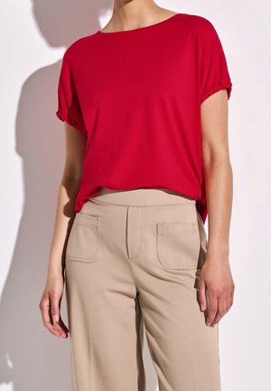 Kobieta nosząca luźną czerwoną bluzkę z krótkim rękawem wpuszczoną w beżowe spodnie z wysokim stanem i przednimi kieszeniami.