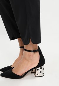 Černé semišové lodičky se špičatou špičkou a stříbrným blokovým podpatkem s puntíky. Disponují řemínkem kolem kotníku s přezkou; měkká textura a strukturovaný design.
