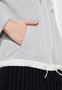 Sudadera con capucha gris claro con un cordón blanco, que presenta un bolsillo delantero y una sección inferior negra sólida con rayas blancas finas.