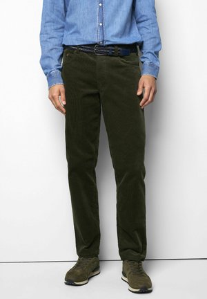 Mann trägt dunkelgrüne Cordhose, navy-blauen gewebten Gürtel, olivgrüne Sneakers und ein hellblaues Hemd mit Knöpfen, steht an einer weißen Wand.