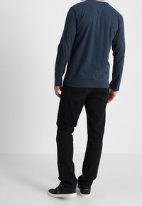 Navyblauw katoenen shirt met lange mouwen, zwarte jeans en donkere sneakers, gezien vanaf de achterkant, waarmee de ronde halslijn en de losse pasvorm van het shirt worden benadrukt.