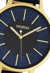 Montre ronde avec un boîtier en or, cadran noir avec des éclats bleus, index et aiguilles dorés. Un bracelet noir complète le design.