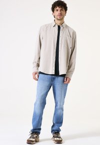 Lichtbeige overhemd met knopen en een zak, gedragen over een zwart T-shirt, gecombineerd met lichtblauwe jeans en bruine sneakers met een patroon.