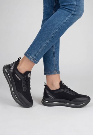 Personne portant des baskets Giesswein noires et un jean bleu sur un fond clair uni, un pied légèrement levé derrière l'autre.
