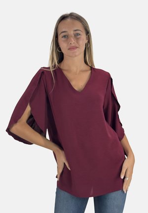 Bordeaux lichtgewicht top met een V-hals en brede mouwen met uitsparingen. Soepele textuur en een iets relaxte pasvorm. Gecombineerd met blauwe jeans.