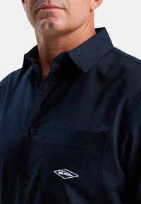 Marineblauwe overhemd met een kraag, voorzak en geborduurd logo. Gemaakt van een gladde stof met een subtiele textuur en contrasterende rode details.