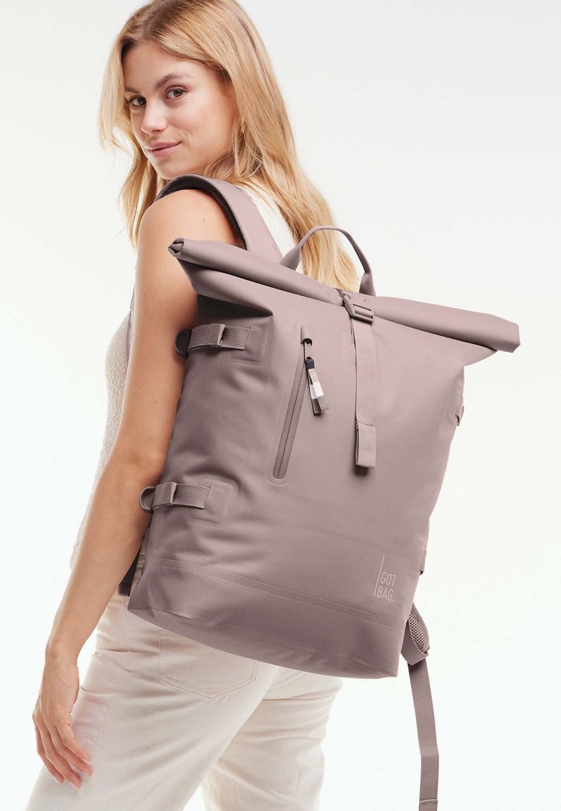 Jeune femme aux longs cheveux blonds portant un grand sac à dos mauve à rabat enroulable et un pantalon clair, regardant par-dessus son épaule.