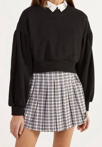Donna che indossa un maglione corto nero con maniche a sbuffo sopra una camicia a colletto, abbinato a una gonna plissettata a quadri bianca e nera.
