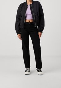 Svart bomberjacka med ribbade ärmslut över en ljuslila croppad topp, ihop med svarta raka jeans och svarta sneakers med vita sulor.