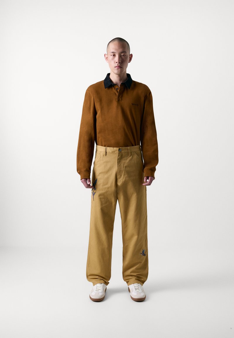Braun langärmliges Poloshirt mit schwarzem Kragen, kombiniert mit beigen weit geschnittenen Hosen mit blauer Stickerei und weißen Sneakers.