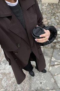 Mörkbrun trenchcoat med stora knappar över en grå tröja. Håller en svart kaffekopp, med en svart flätad handväska. Svarta byxor och skor.