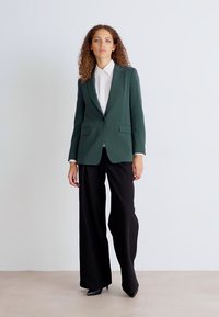 Tmavě zelený blazer se strukturovaným designem, dvěma předními kapsami a zapínáním na jeden knoflík, v kombinaci s černými širokými kalhotami. Pod ním bílá košile.