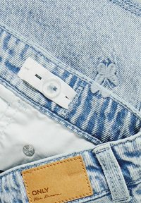 Blå denimtyg med texturerade mönster, vit knapp och spänne, ljusa sömmar och en brun etikett med texten "ONLY Blue Denim."