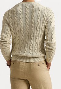 Pull en maille torsadée crème avec un motif texturé, présentant des poignets et un ourlet côtelés. Associé à un pantalon de couleur claire et un subtil détail de poche arrière.