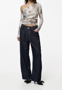 Glänzendes, strukturiertes, silber-graues Oberteil mit einseitigem Schulterdesign, kombiniert mit dunkelblauen, weit geschnittenen Jeans aus Denim mit Knopfverschluss und Taschen.