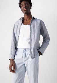 Camisa de botones a rayas azul y blanca sobre una camiseta sin mangas blanca, combinada con pantalones de tiro ajustable azul claro con detalles dorados.