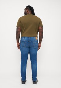 Olivengrønn t-skjorte med korte ermer sammen med blå denimjeans. Jeansene har baklommer og en lys skinnlapp ved livet.