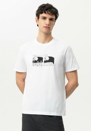 Man met kort donker krullend haar, die een wit T-shirt draagt met een zwart-witte cartoonprint en zwarte pantalon met fijne strepen.