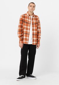 Camisa de franela a cuadros en naranja y blanco con un bolsillo, llevada sobre una camiseta blanca, combinada con pantalones cargo negros y zapatillas negras.