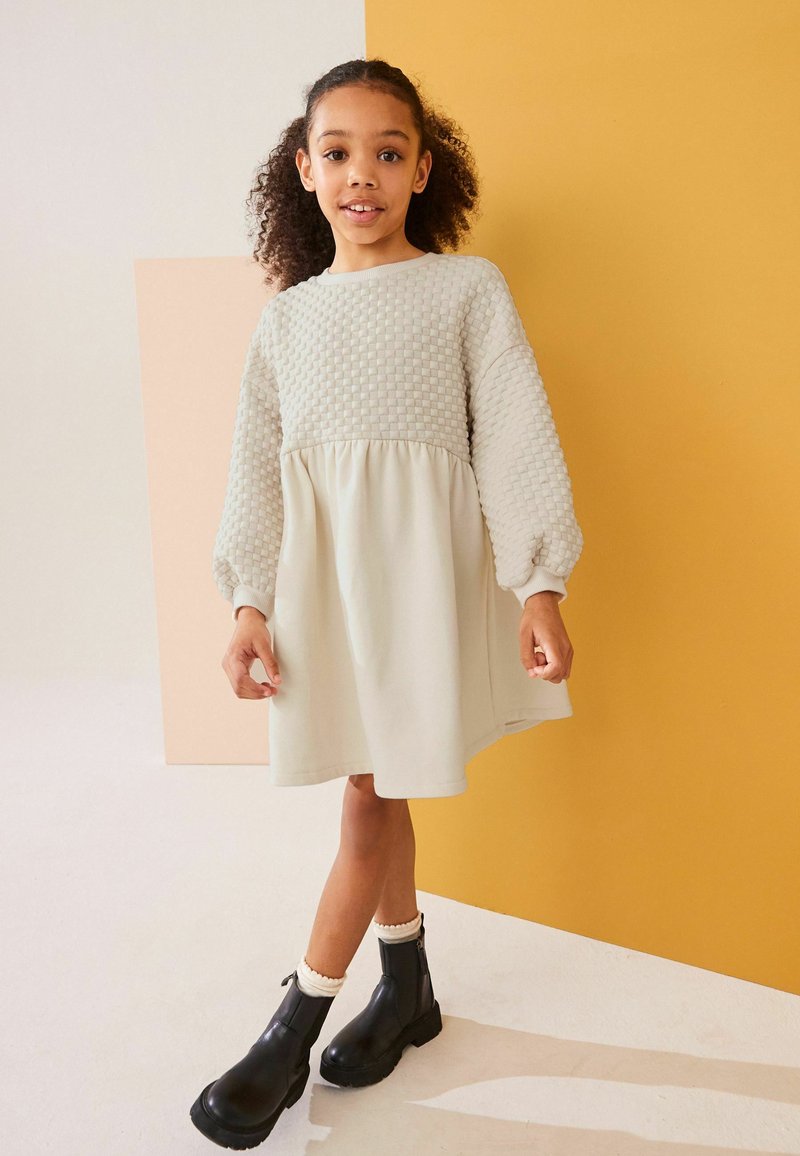 Vaaleanharmaa mekko, jossa on tekstiilistä valmistettu vartalo-osa ja virtaava hame, yhdistettynä mustiin nilkkureihin ja valkoisiin sukkiin, keltaista ja pehmeää vaaleanpunaista taustaa vasten.