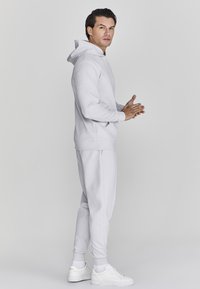 Lichtgrijze hoodie van zacht materiaal, met een voorzak en een capuchon met trekkoord, gecombineerd met bijpassende joggingsbroek en witte sneakers.