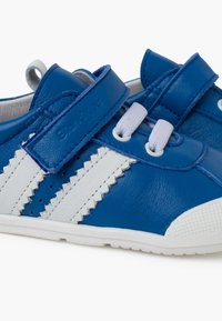 Zapatos infantiles de cuero azul con acentos blancos en zigzag y correas de velcro. Presentan una suela de goma blanca texturizada y detalles de ojal para un cordón decorativo.